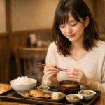 和食屋で定食を前に箸を持つ日本人女性。湯気の立つご飯と味噌汁、焼き魚が並ぶ木目のテーブルと柔らかな店内光が、食事前の静かな期待感を伝える構図。 A Japanese woman holding chopsticks before a teishoku meal, with steam rising from rice and miso soup, captured in warm natural light inside a traditional restaurant.