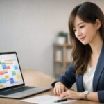 Googleカレンダー風のカラフルな予定表をノートPCで確認しながら、ネイビージャケット姿の若い東アジア系女性がデスクで穏やかにスケジュールを立てている写真。A young East Asian woman in a navy blazer calmly plans her schedule at a light wooden desk while viewing a color-coded Google Calendar–style planner on her laptop.